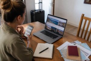 Iemand vergelijkt reisopties op een laptop en smartphone met notities op tafel
